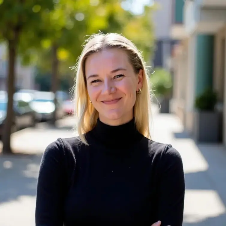 Professional headshot of Sarah Mitchell, a blonde woman in a black turtleneck smiling warmly at the camera in an outdoor urban setting with soft natural lighting.